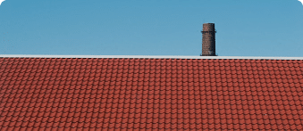 Terracotta roof tiles arranged in neat rows with a weathered brick chimney rising above the roofline against a clear blue sky.