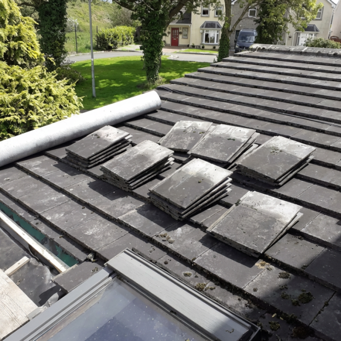 Pitched roof with stacked slate tiles, large roll of underlayment, integrated skylight, and mature trees with house visible in background.