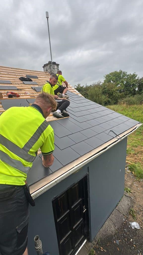 Three roofers in high-visibility vests laying dark roof tiles on a pitched roof beside a grey outbuilding and tall antenna mast.