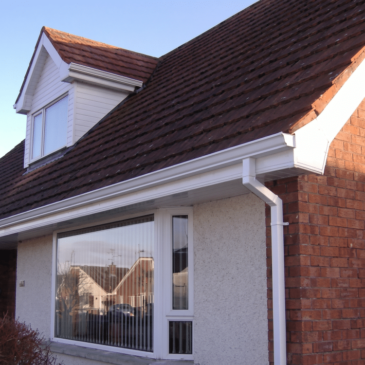 Residential house exterior showing dark brown roof tiles, white uPVC guttering and downpipe, cream plastered wall, red brick section, large window with reflections of neighboring houses and blue sky.