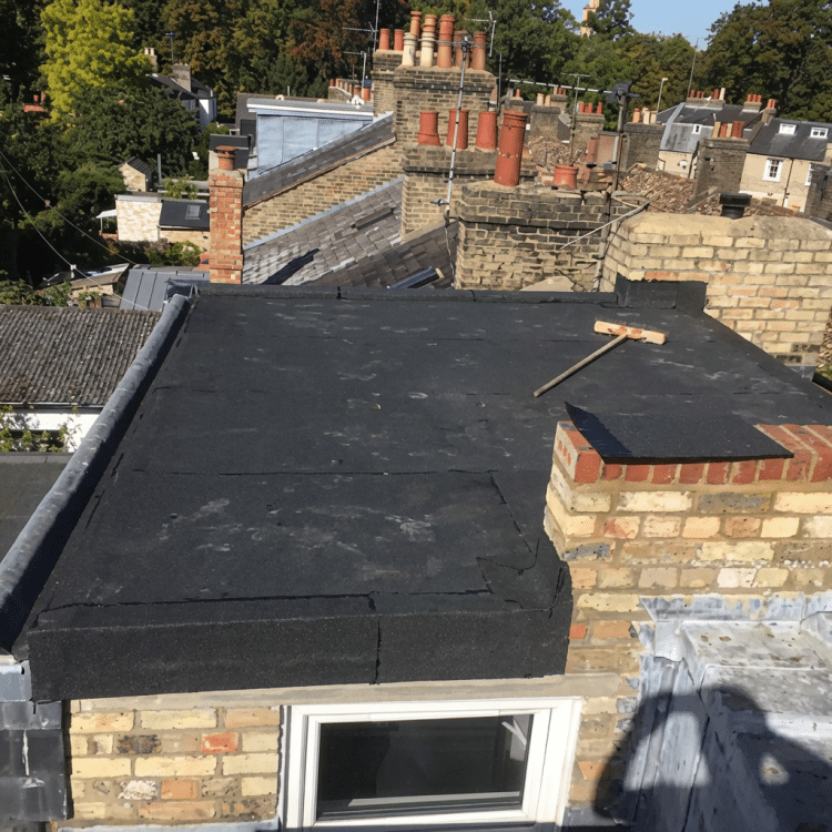 An elevated view of a black flat roof under repair, featuring felt layers and a broom resting on the surface. The roof is surrounded by brick chimneys and neighboring terraced houses in an urban setting.