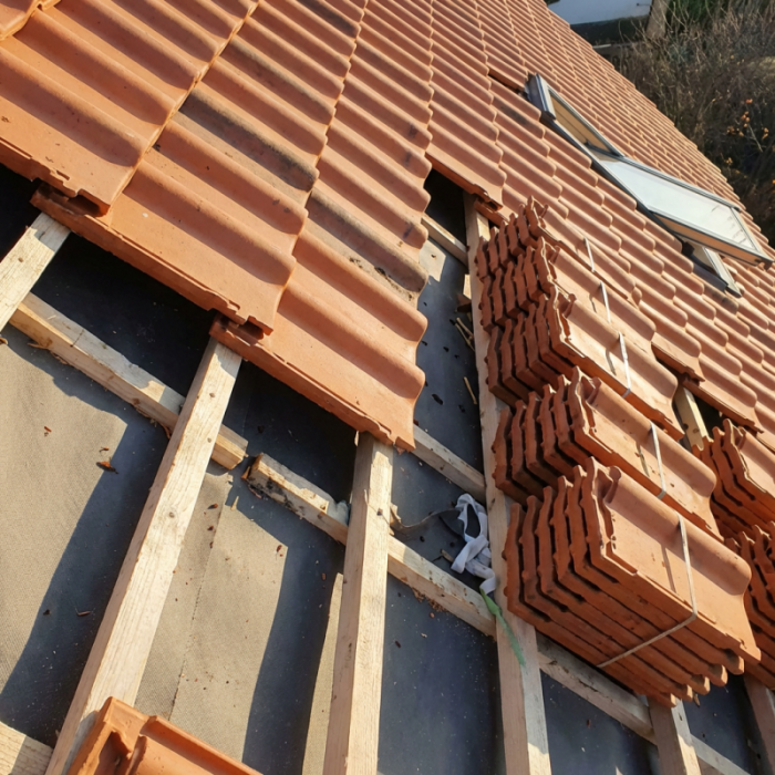 Roof installation in progress with terracotta tiles being laid on wooden battens, stacked tiles on the right, and background vegetation visible.
