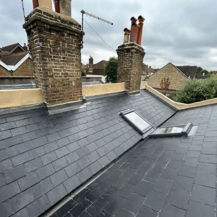 Newly tiled slate roof with two skylights and two brick chimneys under an overcast sky.