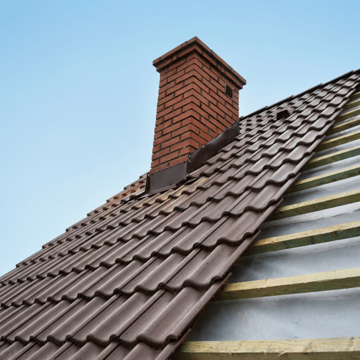 Roof under construction with new dark brown interlocking tiles, exposed wooden beams, silver insulation material, brick chimney, and clear blue sky.