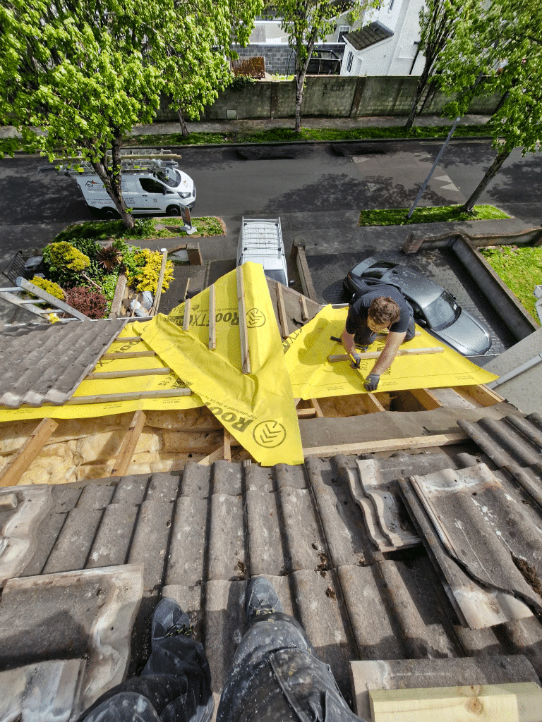 A roofer rolls out yellow membrane on an exposed residential roof section overlooking a suburban street.