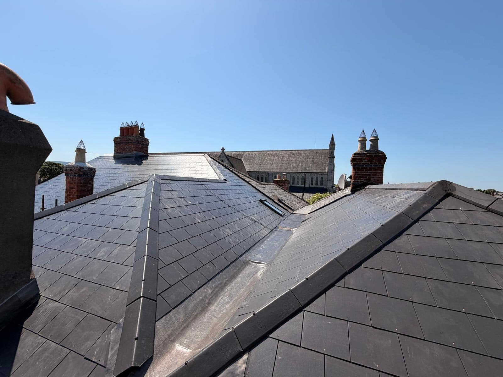 Wide angle view of an intricate, fully tiled slate roof structure with metal-flashed valleys and multiple brick chimneys under a clear sky.